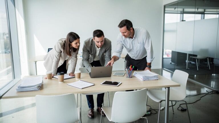 Tri kolege savijene nad laptopom tokom sastanka u svetlom modernom kancelarijskom prostoru